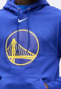 Άτομο που φορά μπλε φούτερ με κουκούλα με κίτρινο κυκλικό λογότυπο της Γέφυρας Γκόλντεν Γκέιτ και λευκό σήμα Nike από πάνω, με τα χέρια στην εμπρός τσέπη.