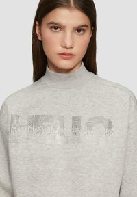 Grijze sweatshirt met hoge kraag, voorzien van een "HELLO"-ontwerp in glanzende strasssteentjes aan de voorkant, gemaakt van zacht materiaal met een ontspannen pasvorm.