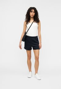 Shorts de sport noirs avec une ceinture élastique, accompagnés d'un débardeur blanc et de baskets blanches. Les accessoires comprennent un petit sac à bandoulière noir.