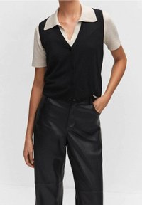 Personne portant une chemise beige à manches courtes avec col sous un gilet noir boutonné, pantalon en cuir noir taille haute, la main dans la poche.