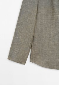 Poignet de manche longue et partie inférieure latérale d'une chemise en lin marron avec un motif à carreaux subtil sur fond blanc.