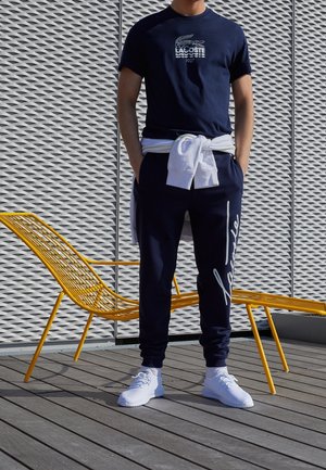 T-shirt bleu marine avec logo Lacoste blanc ; joggers bleu marine avec détails en écriture ; baskets blanches ; chaise en fil jaune et arrière-plan gris texturé.