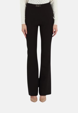 Donna che indossa pantaloni neri a vita alta a zampa con piccolo dettaglio di fibbia argentata, abbinati a tacchi beige a punta e una maglia bianca a fantasia.