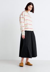 Stripet langermet topp med røde og gule striper, kombinert med et svart, flytende skjørt og brune slip-on sko. Enkelt og avslappet design.