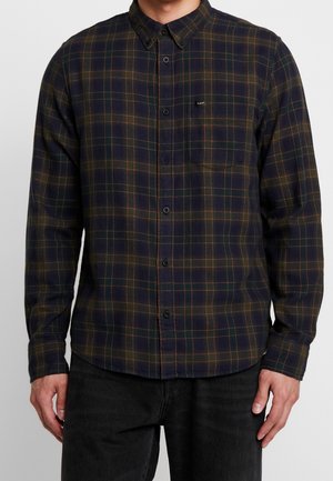 Mann trägt ein langärmeliges, dunkelblau-grün kariertes Hemd mit Brusttasche und schwarze Jeans, steht vor einem einfarbigen Hintergrund.