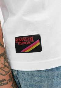 Biała bawełniana koszulka z czarną prostokątną naszywką. Na naszywce widnieje napis "STRANGER THINGS" na czerwono, z różowymi i żółtymi ukośnymi paskami.