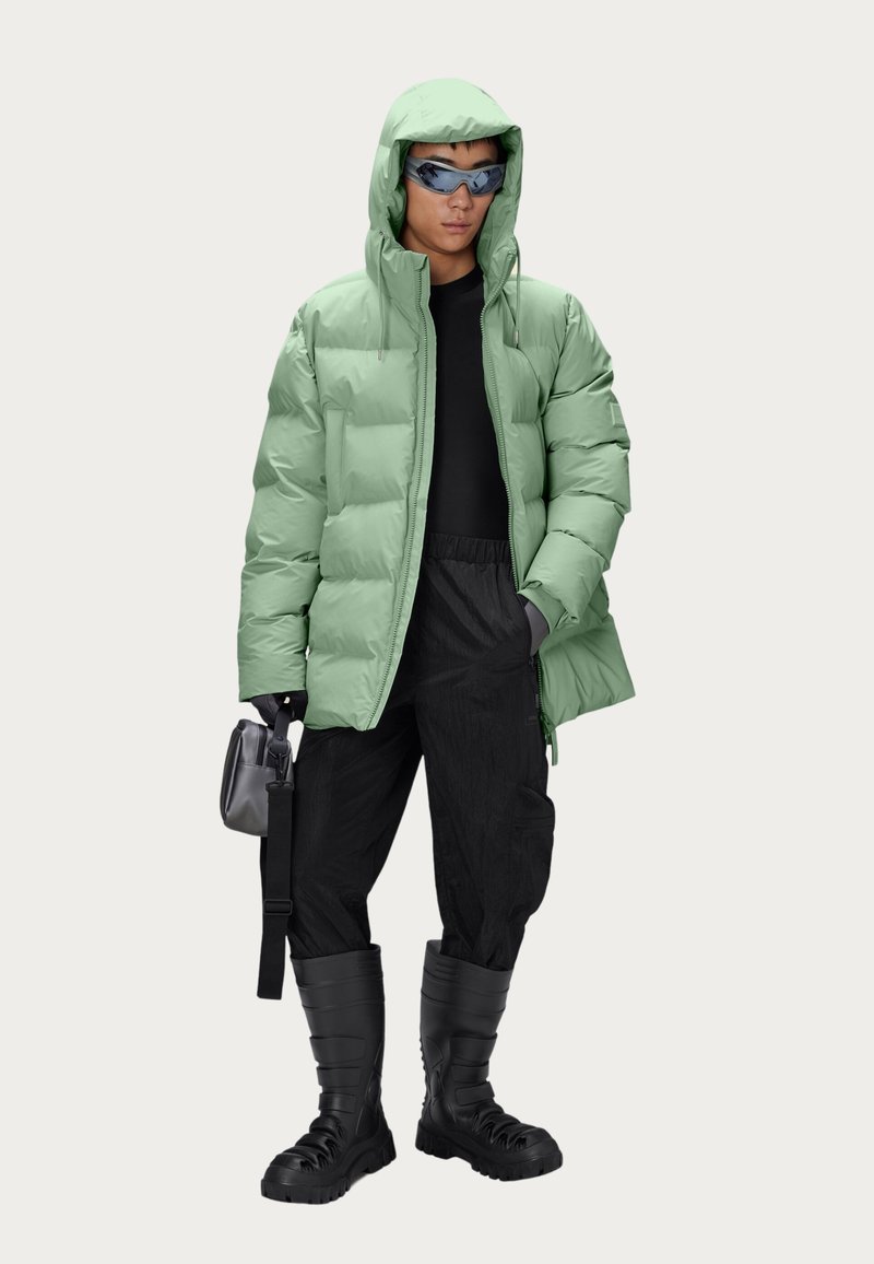 Doudoune vert clair avec capuche, associée à un pantalon noir texturé et des bottes noires montantes. Les accessoires comprennent des lunettes de soleil teintées et un sac.