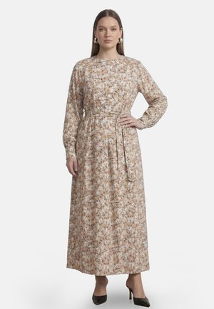 Kvinde med langt hår iført guldringe øreringe og en langærmet blomster maxi kjole med en indsnævret talje og sorte høje hæle.