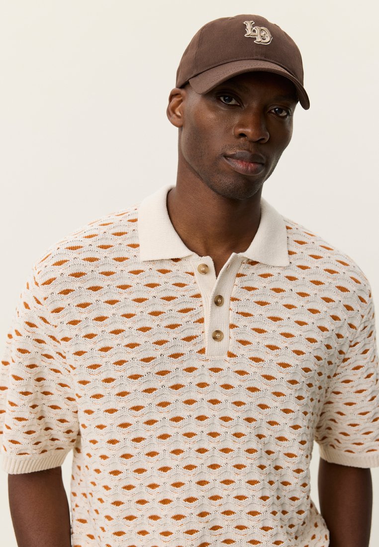 Hellbeiges Strick-Poloshirt mit orangefarbenem Lochmuster, ausgestattet mit einem cremefarbenen Kragen, einer Knopfleiste mit drei Knöpfen und kurzen Ärmeln. Braune Kappe mit Logo.