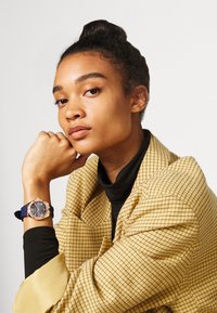 Zlatý hodinky s modrým silikonovým páskem, kulatým ciferníkem, tmavým podkladem a zlatými akcenty. Modelka nosí žlutý kostkovaný blejzr a černý rolák.