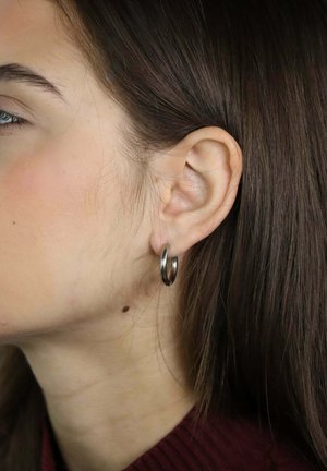 Zbliżenie na lewe ucho kobiety z srebrną kolczykiem kołowym, brązowe włosy wsunięte za ucho oraz mała pieprzyk na szyi.