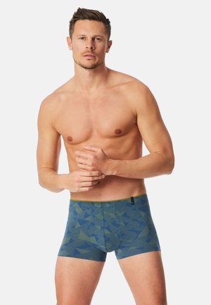 Oberkörperfreier Mann steht mit gefalteten Händen, trägt blaue und grüne gemusterte Boxershorts vor neutralem Hintergrund.