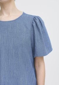 Top din denim albastru, cu mâneci scurte puff, decolteu rotund și linii texturate verticale din fir metalic. Detaliu de design casual subtil.