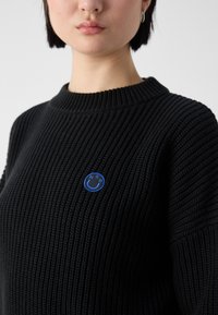 Femme portant un pull côtelé noir avec une petite étiquette bleue en forme de smiley affichant les lettres H, G et O sur la poitrine.