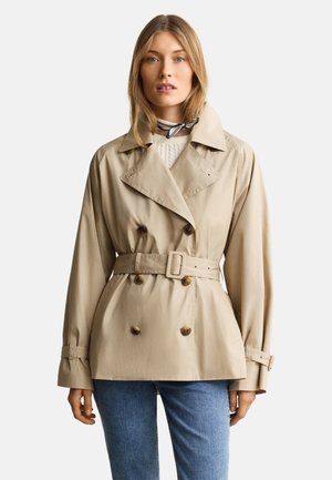 Femme portant un trench-coat beige ceinturé sur un pull blanc et un jean bleu, avec une écharpe rayée autour du cou sur un fond uni.