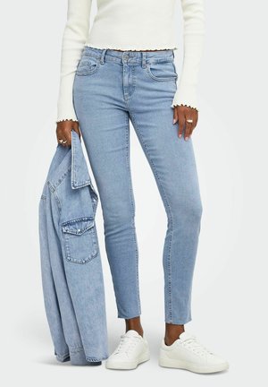 Personne portant un jean skinny bleu clair, un haut blanc à manches longues, des baskets blanches, tenant une veste en denim bleu clair assortie à ses côtés.