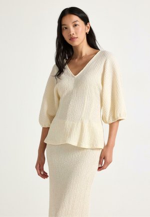 Kvinde med langt sort hår iført en off-white struktureret bluse med V-hals og puffede ærmer samt en matchende nederdel mod en ensfarvet baggrund.