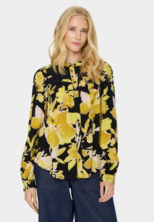 Svart blus med långa ärmar, prydd med ljusgul och rosa blommönster, knäppning fram och plisserade detaljer. Lätt material med mjuk textur.