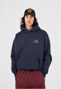 Sudadera azul marino con un bolsillo tipo canguro, con un logotipo bordado en oro. Combinada con pantalones burdeos, el modelo tiene el cabello rizado y oscuro.