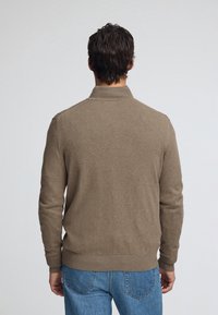 Brun stickad tröja med hög krage, åtsittande mudd och midjeband, bärs över blå jeans. Mjuk textur med minimala synliga sömmar.