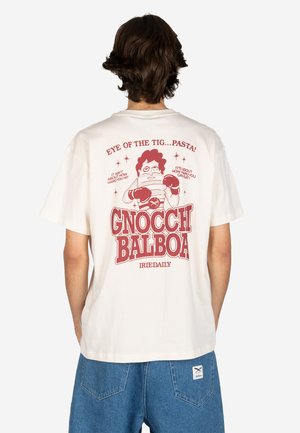 Person trägt weißes T-Shirt mit rotem Cartoon-Boxer-Nudel-Charakter und dem Text „Gnocchi Balboa“ sowie Jeans-Shorts, von hinten gezeigt.