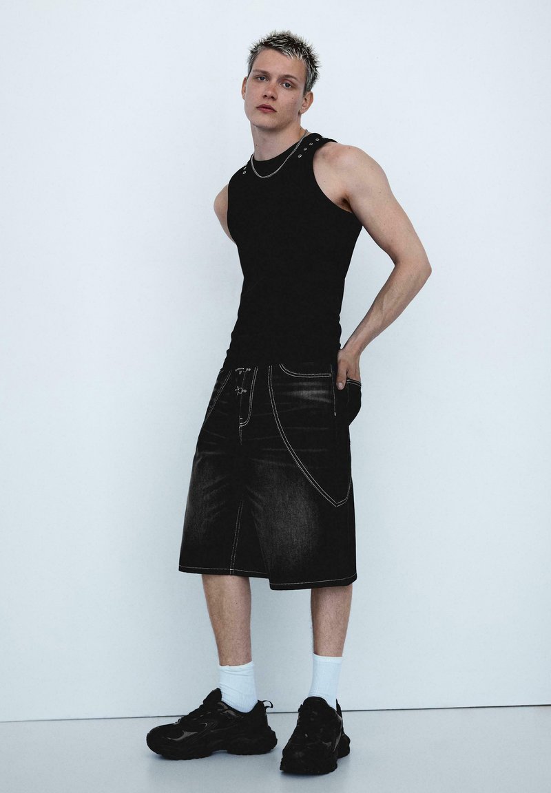 Jeune homme aux cheveux courts décolorés portant un haut noir sans manches, un short en jean noir ample, des chaussettes blanches et des baskets noires, debout contre un mur blanc.