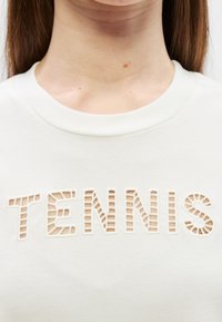 Sweatshirt blanc avec texte découpé "TENNIS" en relief. Tissu doux, design à col rond et coupe décontractée.