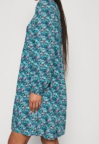 Blommig klänning med ett livligt blått och grönt mönster på en svart bakgrund, långa ärmar, en avslappnad passform och en rund halsringning.