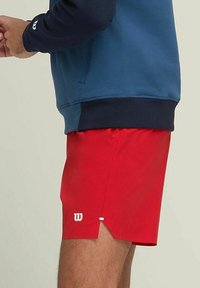 Hombre vestido con pantalones cortos deportivos rojos y un sudadero azul, mostrado desde la cintura hacia abajo sobre un fondo liso.