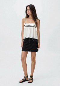 Top bianco senza maniche con bordi scallop neri, abbinato a una gonna di jeans nera con borchie argentate e una silhouette aderente. Sandali neri con cinturini.