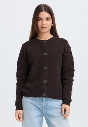 Donna con capelli lunghi e castani che indossa un cardigan marrone scuro a trama con bottoni e jeans azzurri, in piedi davanti a uno sfondo semplice.