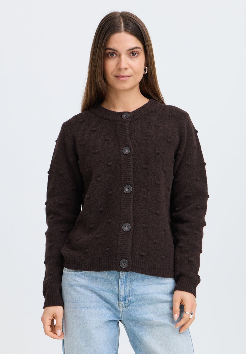 Femme aux longs cheveux bruns portant un cardigan texturé marron foncé boutonné et un jean bleu clair, debout devant un fond uni.