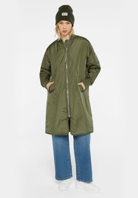 Olivgrüne, langärmelige Jacke mit Reißverschluss, zwei seitlichen Taschen und elastischen Bündchen, getragen mit lockeren blauen Jeans und weißen Sneakers, ergänzt durch eine grüne Mütze.