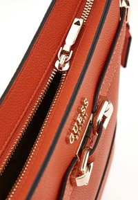 Borsa sintetica arancione con texture, hardware dorato, doppi zip e logo prominente. Presenta una forma strutturata e un bordo nero a contrasto.