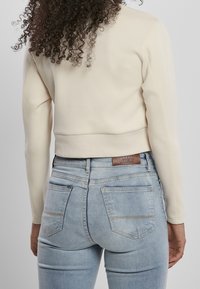 Sudadera corta crema con mangas largas; textura suave. Combinada con vaqueros de mezclilla azul claro que llevan una etiqueta de cuero Urban Classics en la parte trasera.