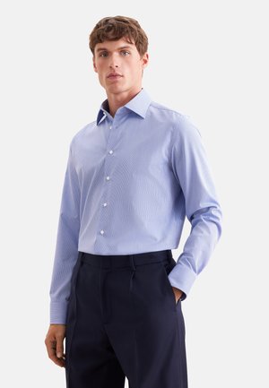 Homme portant une chemise à manches longues bleu clair rayée rentrée dans un pantalon bleu marine foncé, debout avec une main dans la poche.