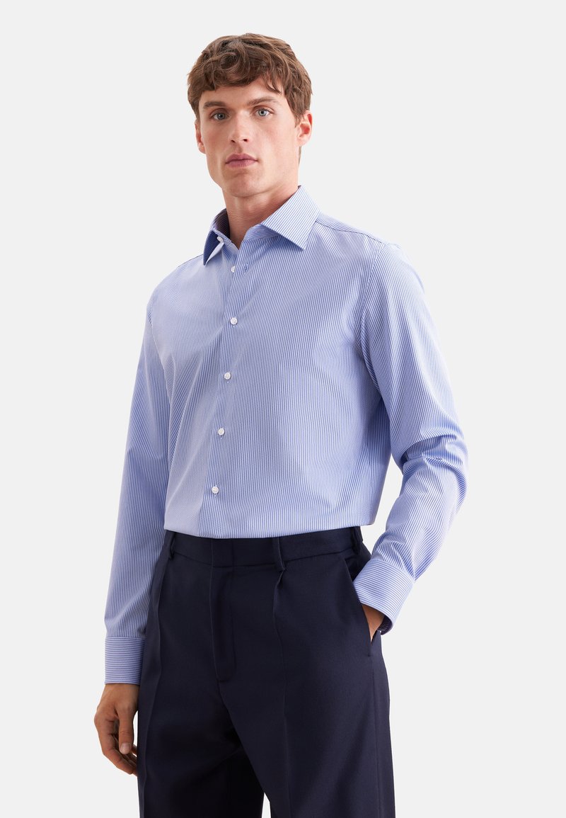 Homme portant une chemise à manches longues bleu clair rayée rentrée dans un pantalon bleu marine foncé, debout avec une main dans la poche.
