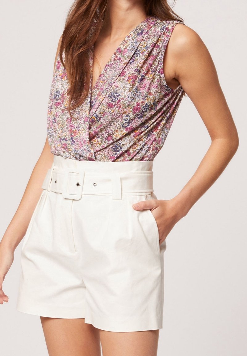 Femme portant un top cache-cœur fleuri sans manches rentré dans un short blanc taille haute avec ceinture, main dans la poche, sur fond uni.