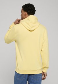 Gul huvtröja av mjukt material med framficka i kängurstil och muddade ärmslut, kombinerad med blå jeans.