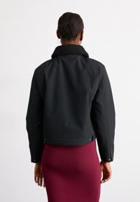 Zwarte cropped jas met een getextureerde shearlingkraag, gladde stof en drukknoopsluitingen. Model draagt een aansluitende bordeauxrode jurk.