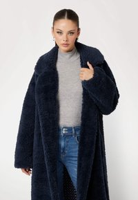 Mørkeblå fluffy frakke med bred krave og lange ærmer, båret over en grå turtleneck og blå jeans, der viser en blød tekstur.