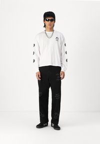 White long-sleeve shirt with black logo and circular graphics, paired with black pants featuring embroidered designs and text, worn with black sneakers.