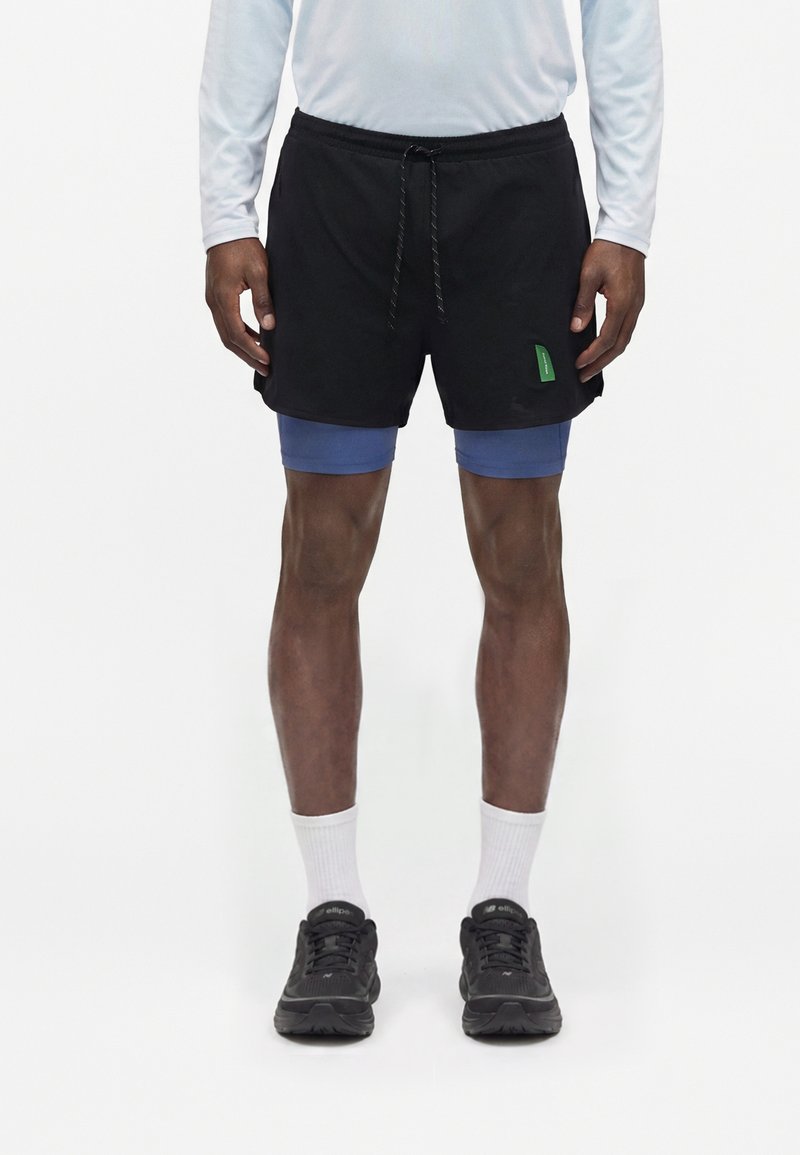 Personne portant un short de sport noir avec un short de compression bleu en dessous, des chaussettes blanches, des chaussures de course noires et un haut blanc à manches longues.
