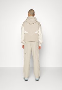 Felpa beige con cappuccio e maniche crema a contrasto, con un disegno geometrico sulla schiena. Abbinata a pantaloni cargo beige chiaro e scarpe blu.