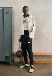 Weiße, flauschige Jacke mit schwarzen Streifen, schwarze Sporthosen mit Logo und weiße Schuhe. Hält einen braunen Basketball in einer Netztasche.