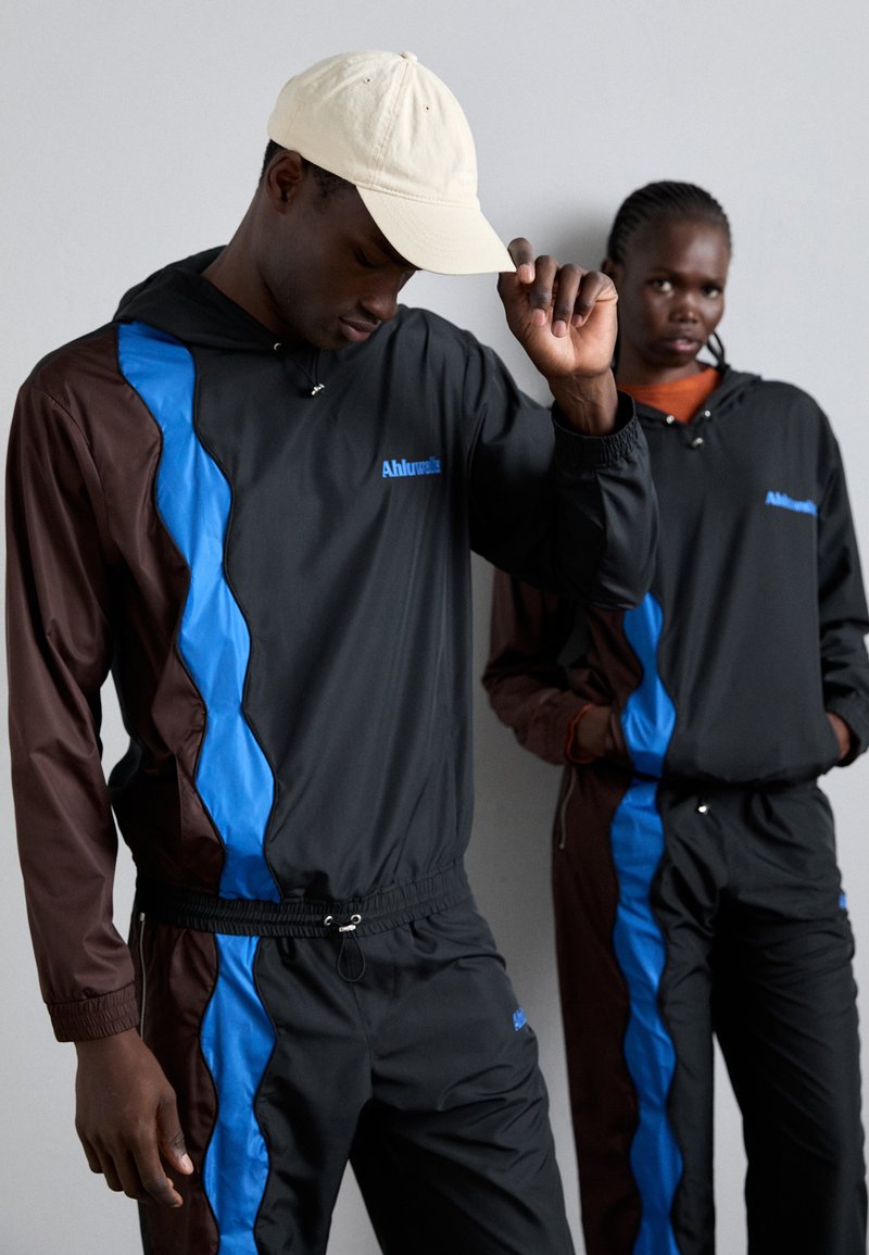 Des survêtements noirs et marron avec des accents bleus, comportant une capuche et des poignets élastiques. Une casquette beige est portée, tandis qu'un deuxième modèle se tient en arrière-plan.
