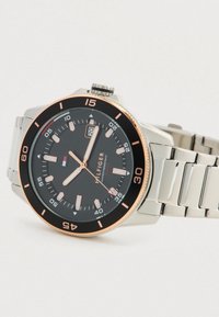 Tommy Hilfiger armbåndsur med sort urtavle, rosegold detaljer, datovisning og sølvfarvet metal kæde, vinklet på hvid baggrund.