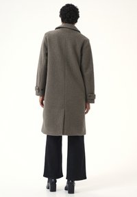 Olijfgroene gestructureerde jas met een notched kraag, lange mouwen en riemmanchetten, gepresenteerd met zwarte wijde broek en hoge laarzen.