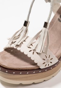 Beige plattform sandal med utskuren blommönster och krämfärgade tofsar. Har silverdetaljer och en strukturerad sula med scallopkant.