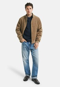 Uomo che indossa una giacca in velluto a coste marrone sopra un maglione blu navy, con jeans blu chiaro e scarpe in pelle marrone scuro, in piedi con una mano in tasca.